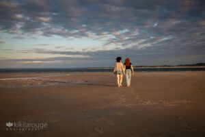 Mom and redheaded daughter walking away on sandy beach with dramatic sunset skies