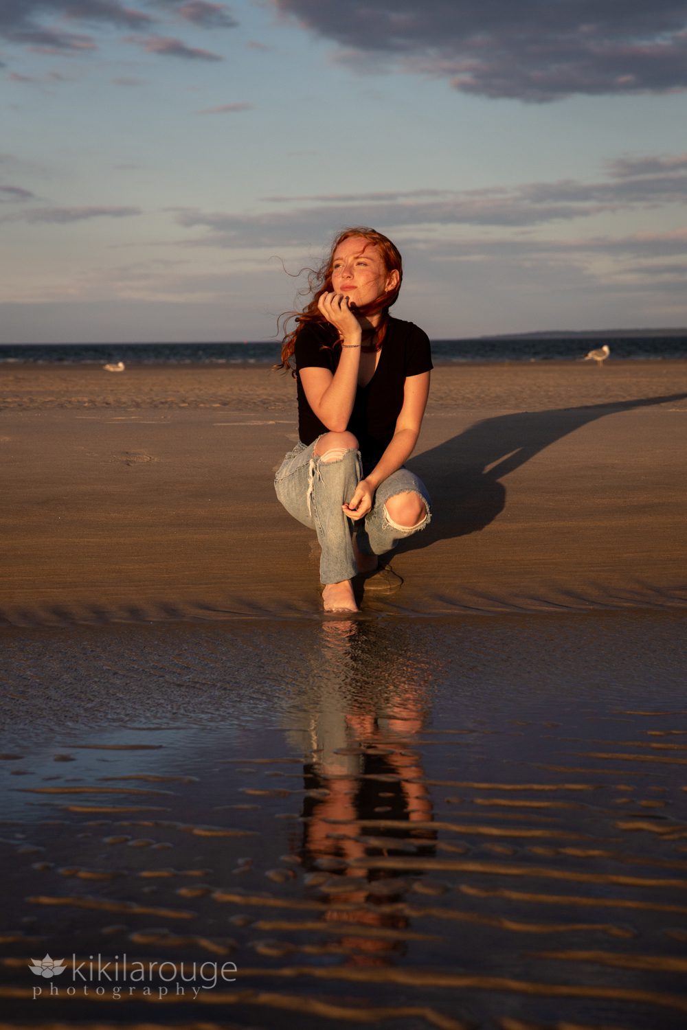 Redhead in ripped jeans black top by tidepool with reflection looking off at sunset hair blowing
