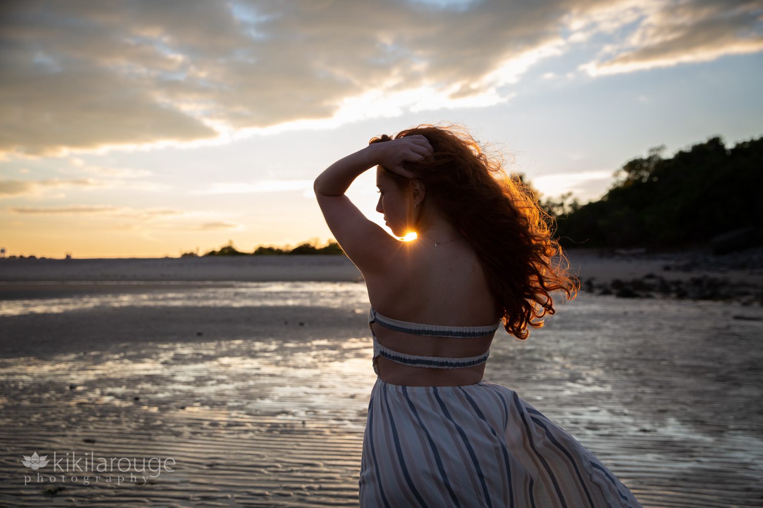 Redhead profile at sunset with dress blowing and had on head and sun peeking through by neck