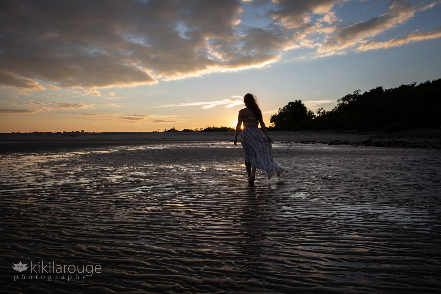Silhouette of girl with blowing dress walking at low tide towards sunset