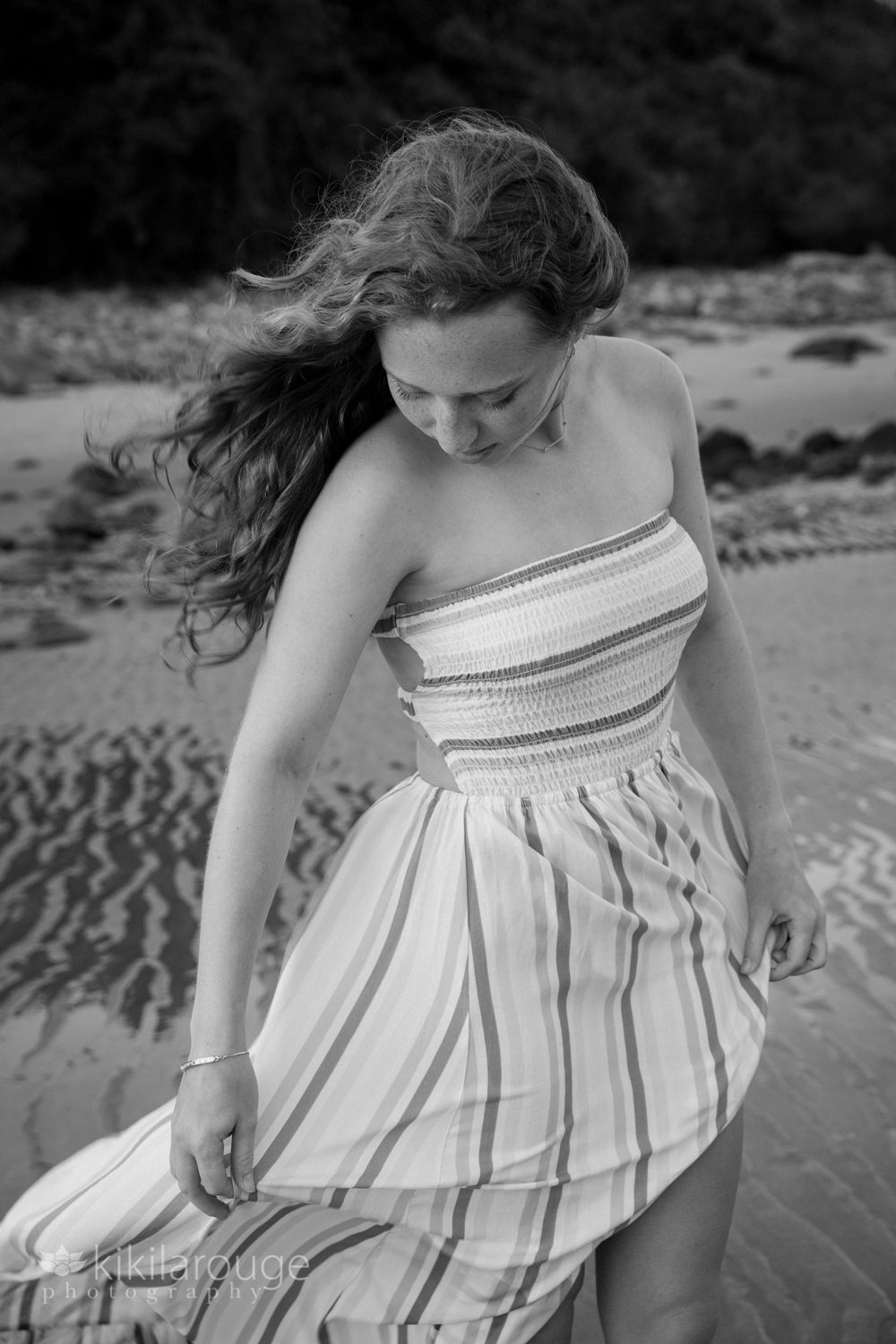BW of girl with hair blowing at beach looking down holding flowing dress