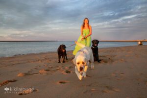 Girl in with long blonde hair holding three dogs on leaches on beach at sunset