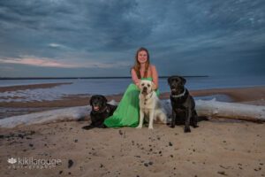 Teen girl in long green dress sitting on driftwood at beach with three dogs