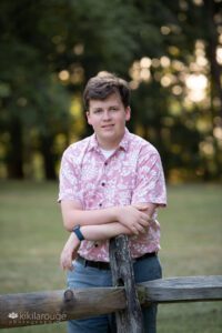 Teen boy brown hair pink shirt green shorts leaning on wooden fence post
