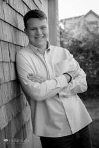 Senior boy portrait in buttons down shirt and jeans leaning on weathered shed