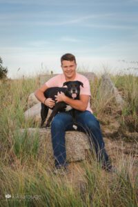 Boy in peach tee and jeans sitting on rock at dune beach with his cute Pittie dog