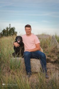 Boy in peach tee and jeans sitting on rock at dune beach with his black lab dog