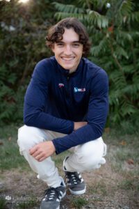 Teen boy squatting down smiling by green bush in white sweatpants