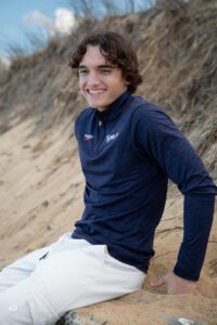 Teen boy with curly brown hair in blue top sitting at beach laughing
