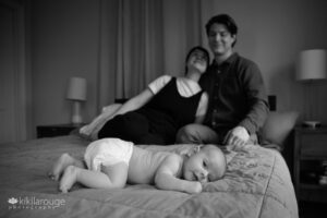 Two month baby in white diapers in foreground with parents smiling in backdrop