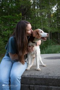Girl with long brunette hair hugging and kissing her dog whose looking off into sunset