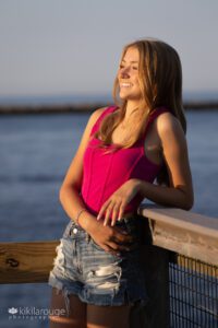 Teen blonde in hot pink top and jeans shorts at beach end of boardwalk overlooking water smiling into the sunset