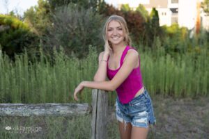 Blonde teen with hot pink shirt and jean shorts leaning on weathered fence by beach dunes smiling