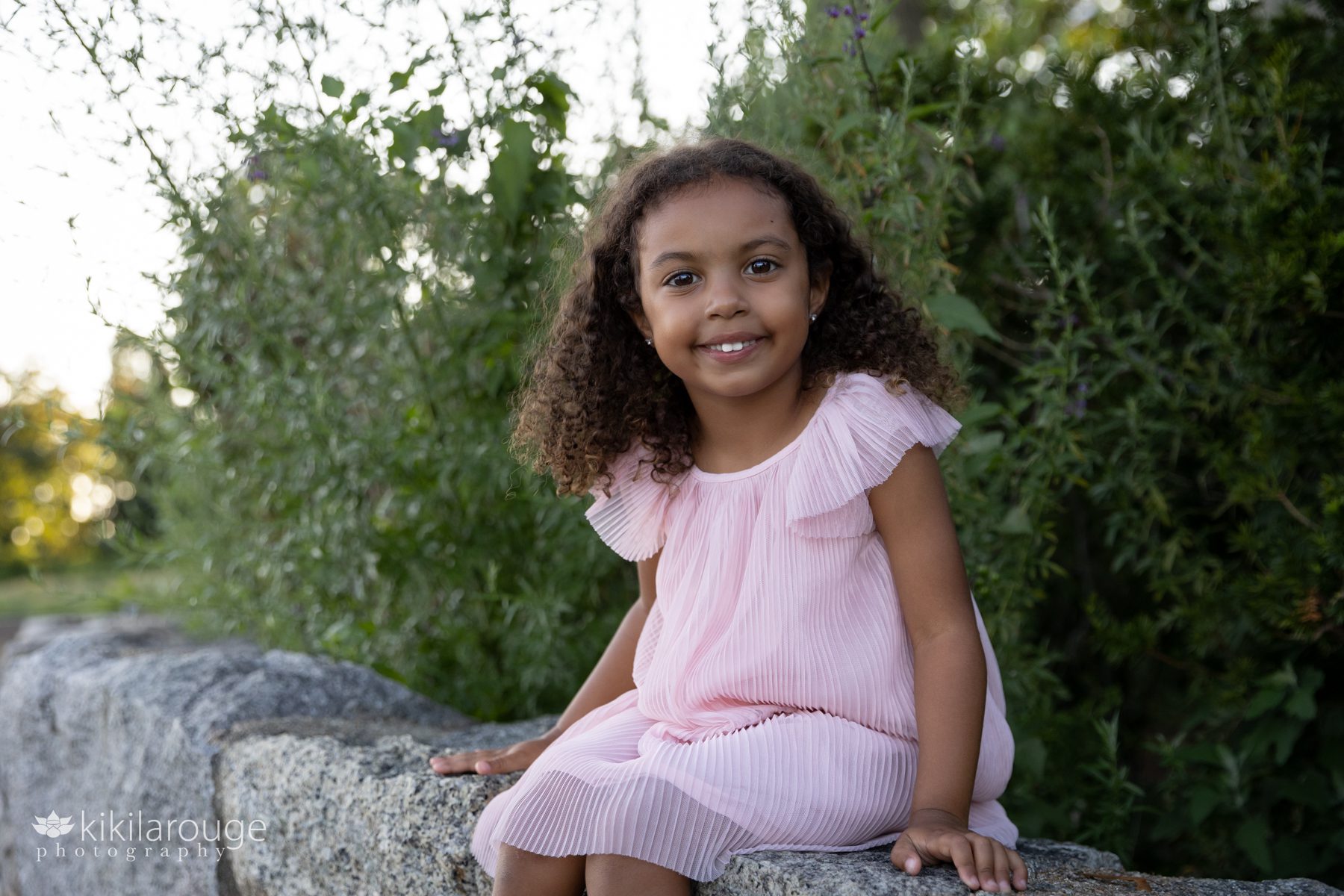 Adorable little girl with black curly hair sitting on rock wall in pink dress