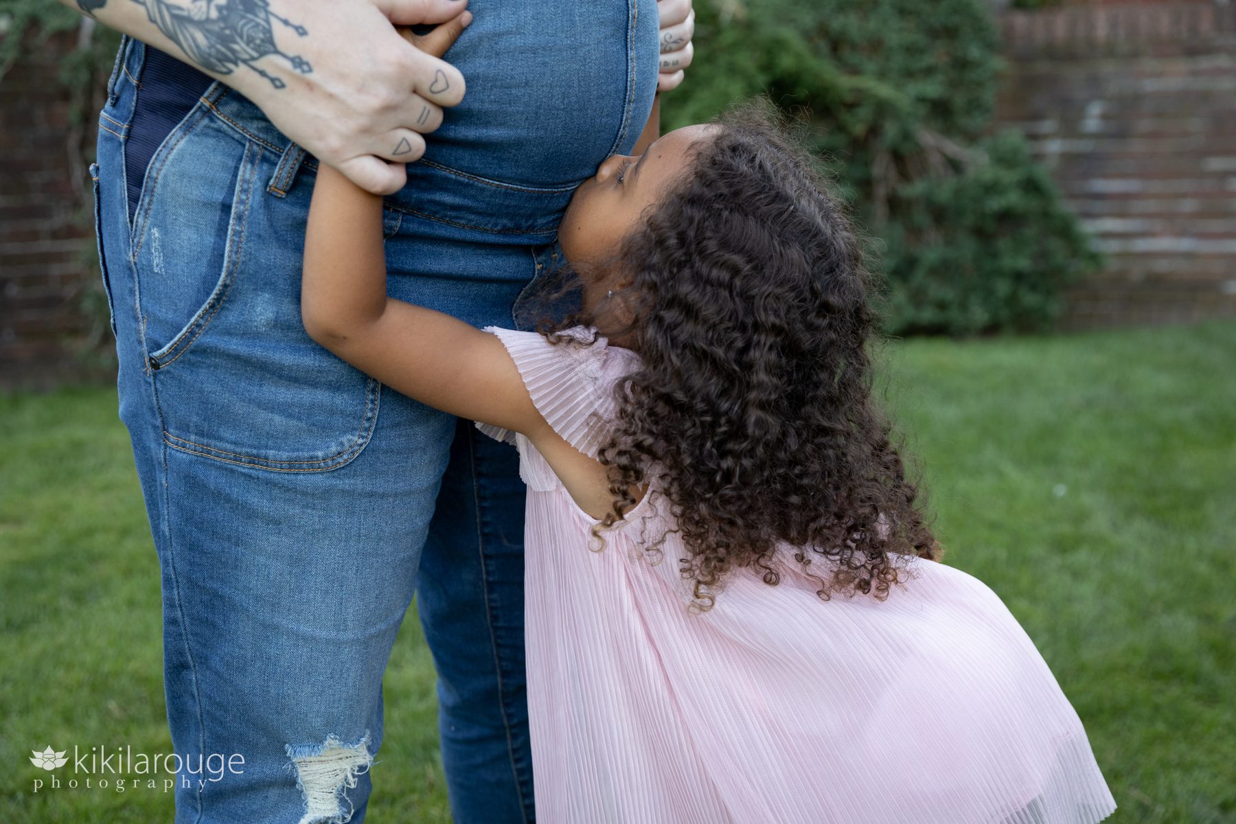 Little girl in blowing pink dress kissing her Mom's pregnant belly