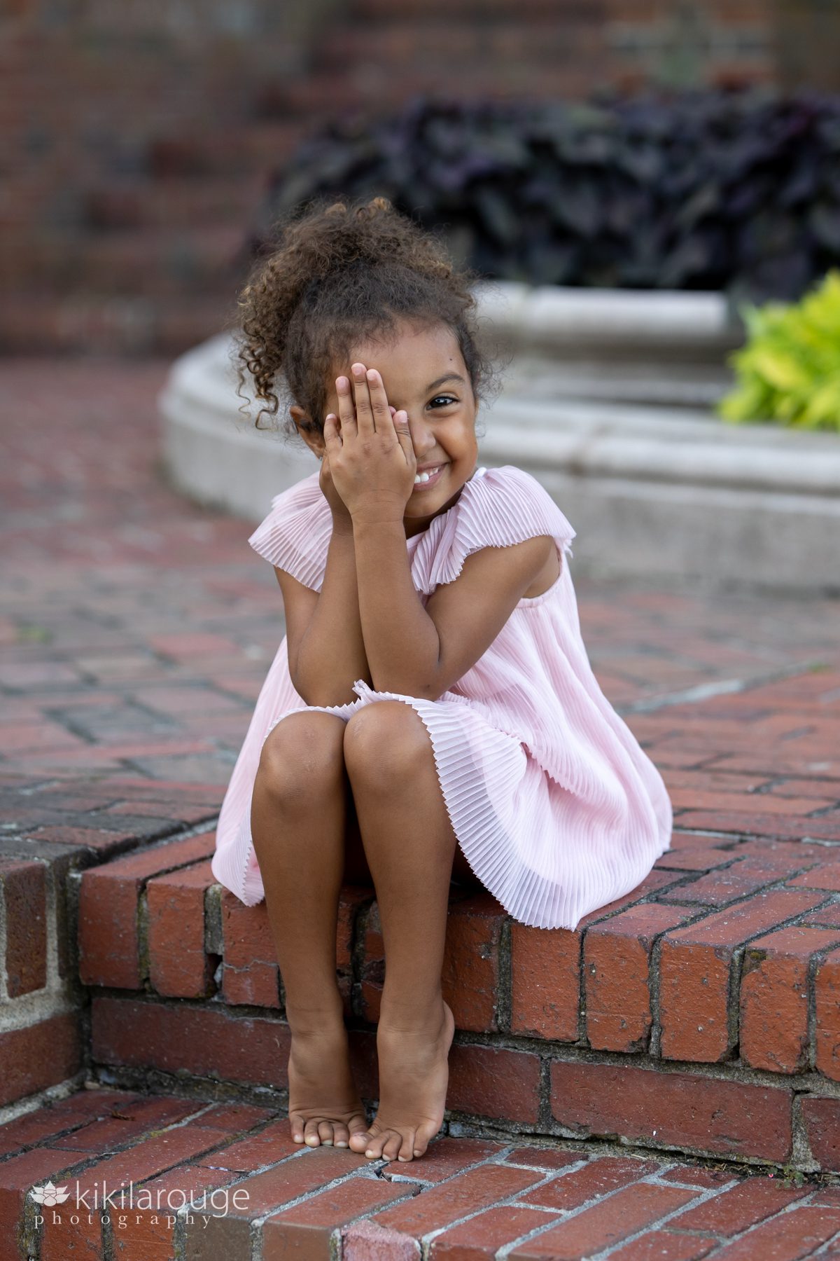 Little girl sitting on brick steps smiling with one eye covered with her hand