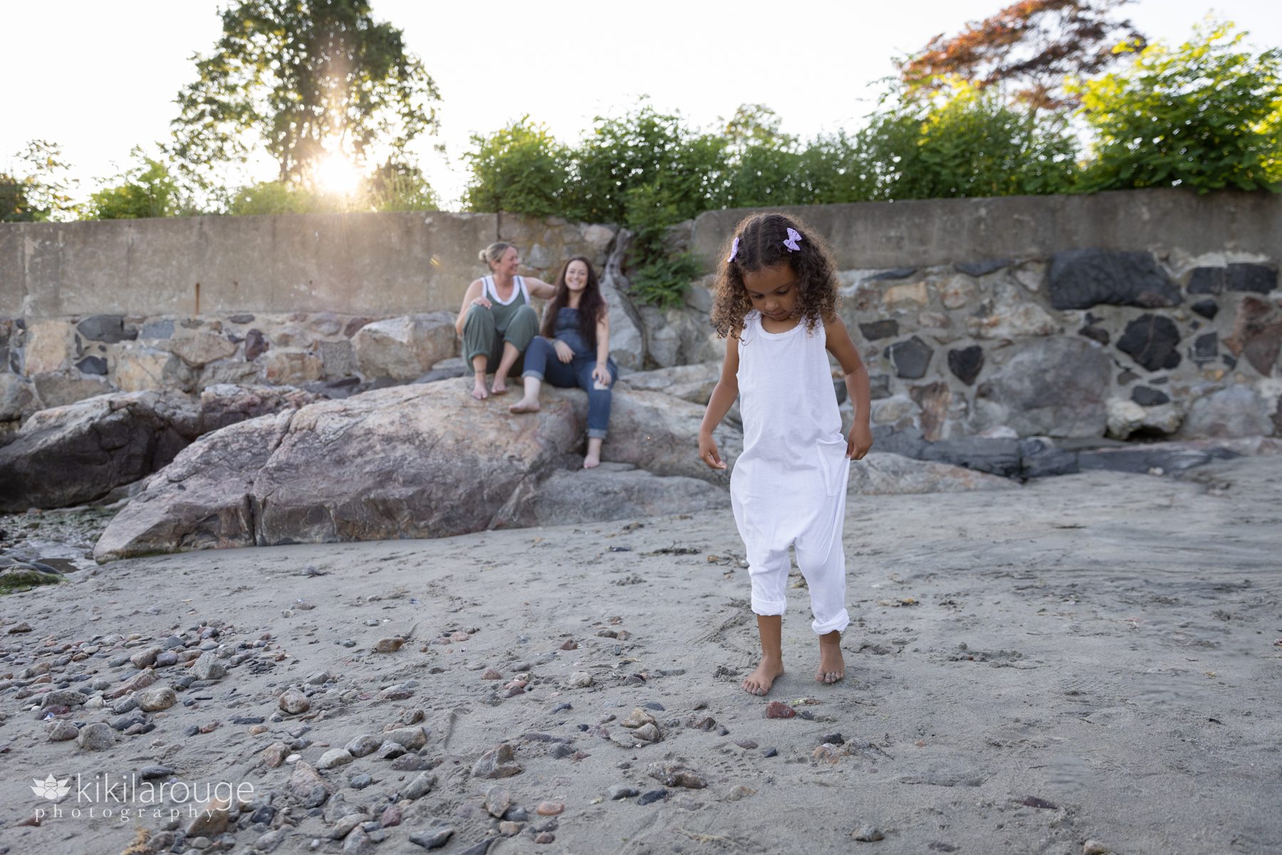 Cute little girl in white jumper looking down with parents sitting on rock at beach in backdrop
