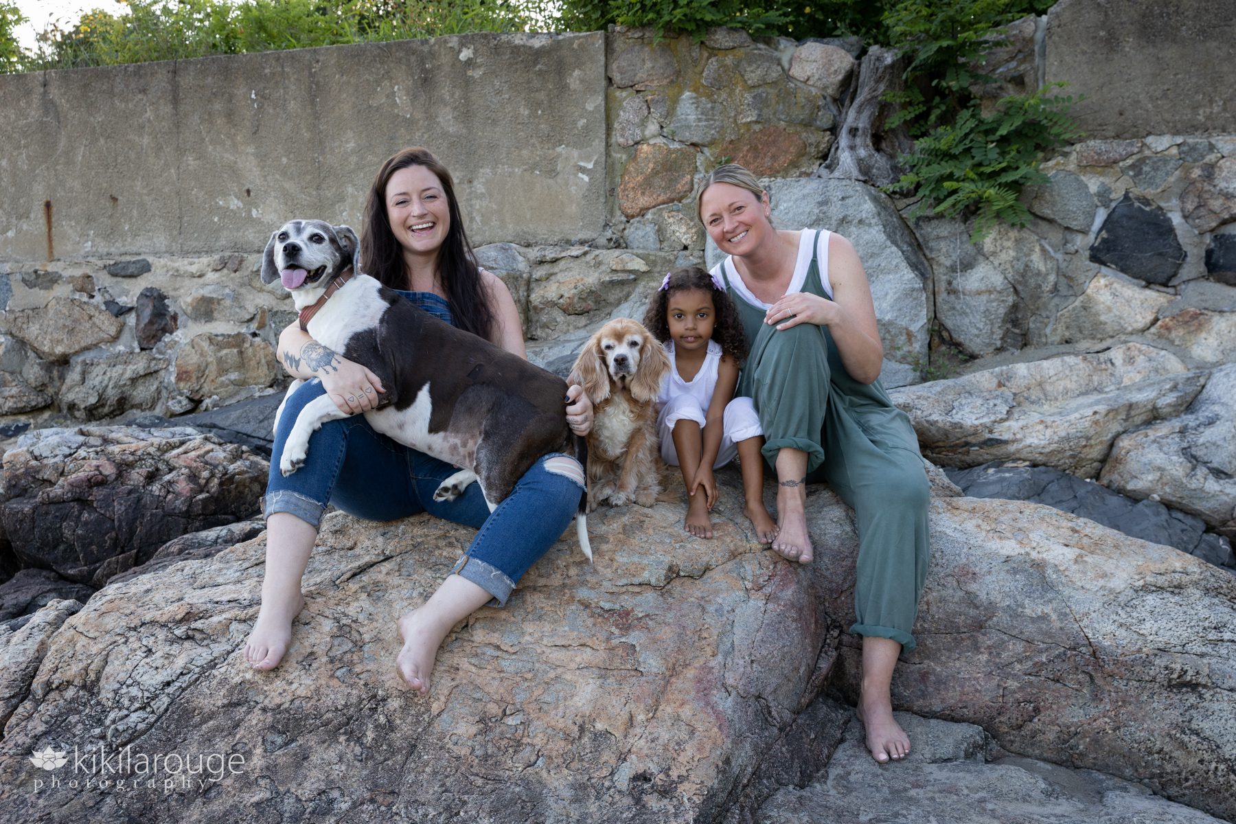 Family photo with two Moms, cute little five year old girl and two Senior Rescue Dogs sitting on beach rocks