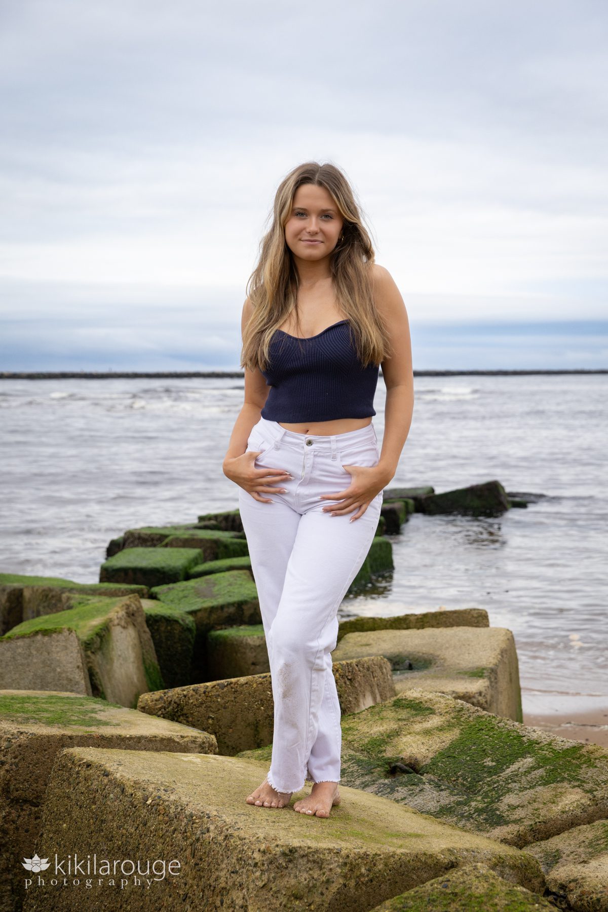 Teen girl white jeans blue top standing on seaweed covered blocks at Plum Island beach
