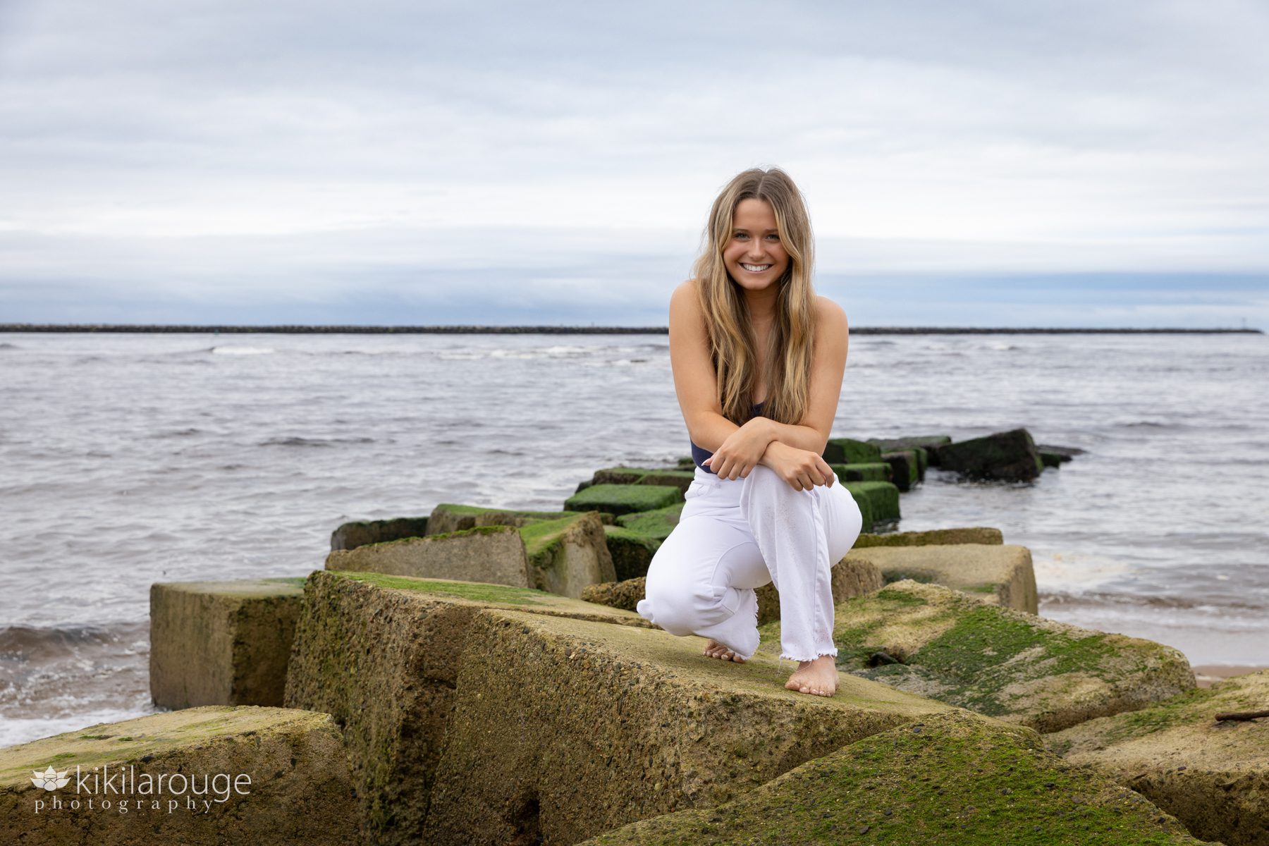 Teen girl white jeans blue top crouching down on seaweed covered blocks at Plum Island beach