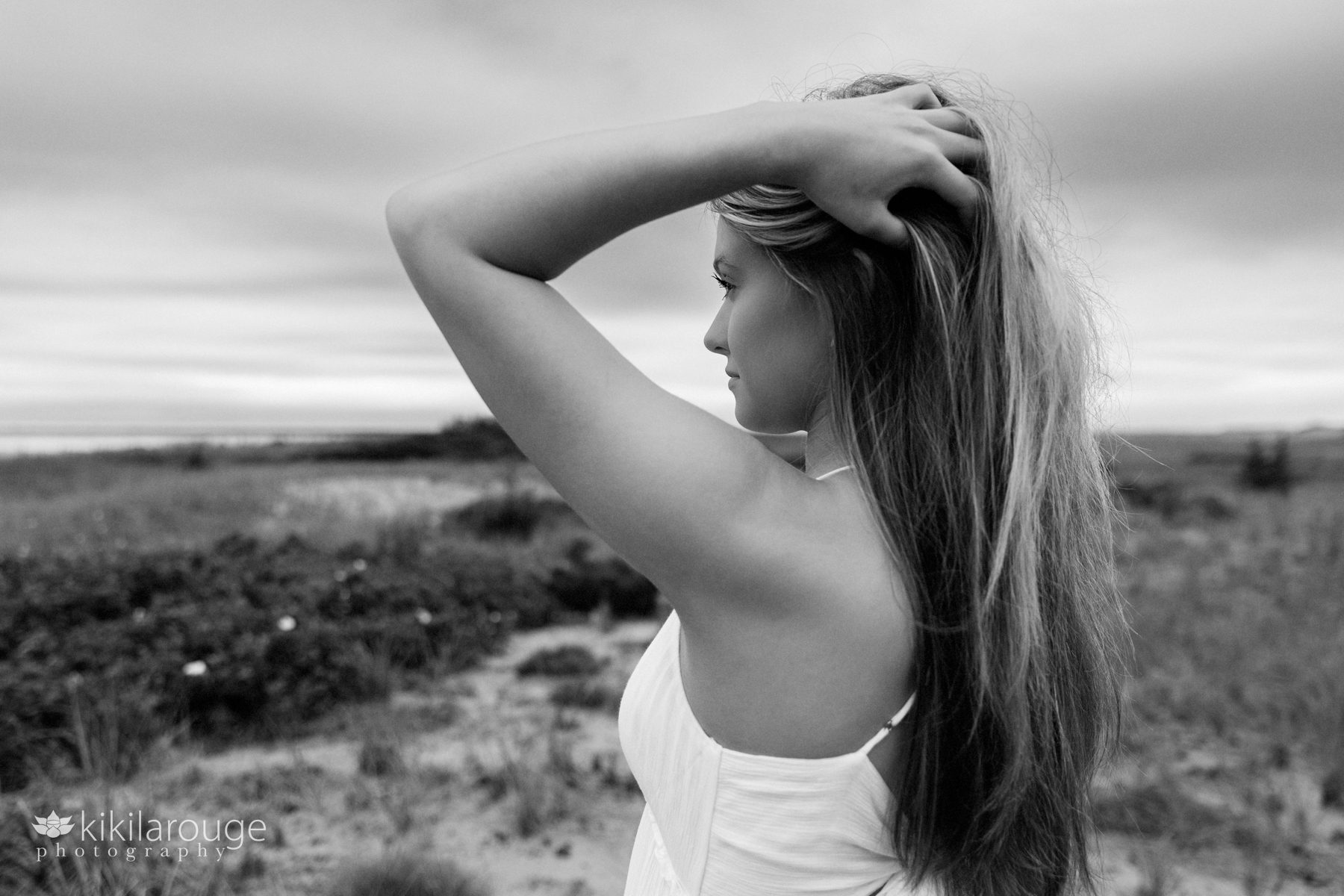 Senior girl long blonde hair with hands on head looking out at dunes and ocean