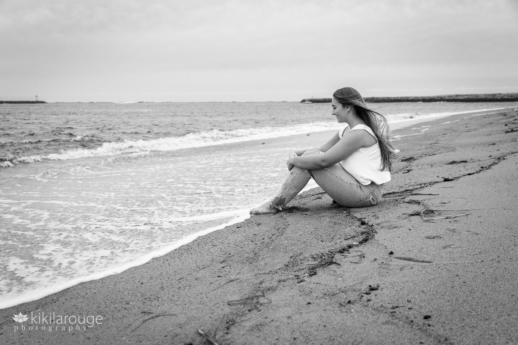 Triton Senior Portrait Girl in Jeans with white top tied in back at beach sitting at water's edge in jeans