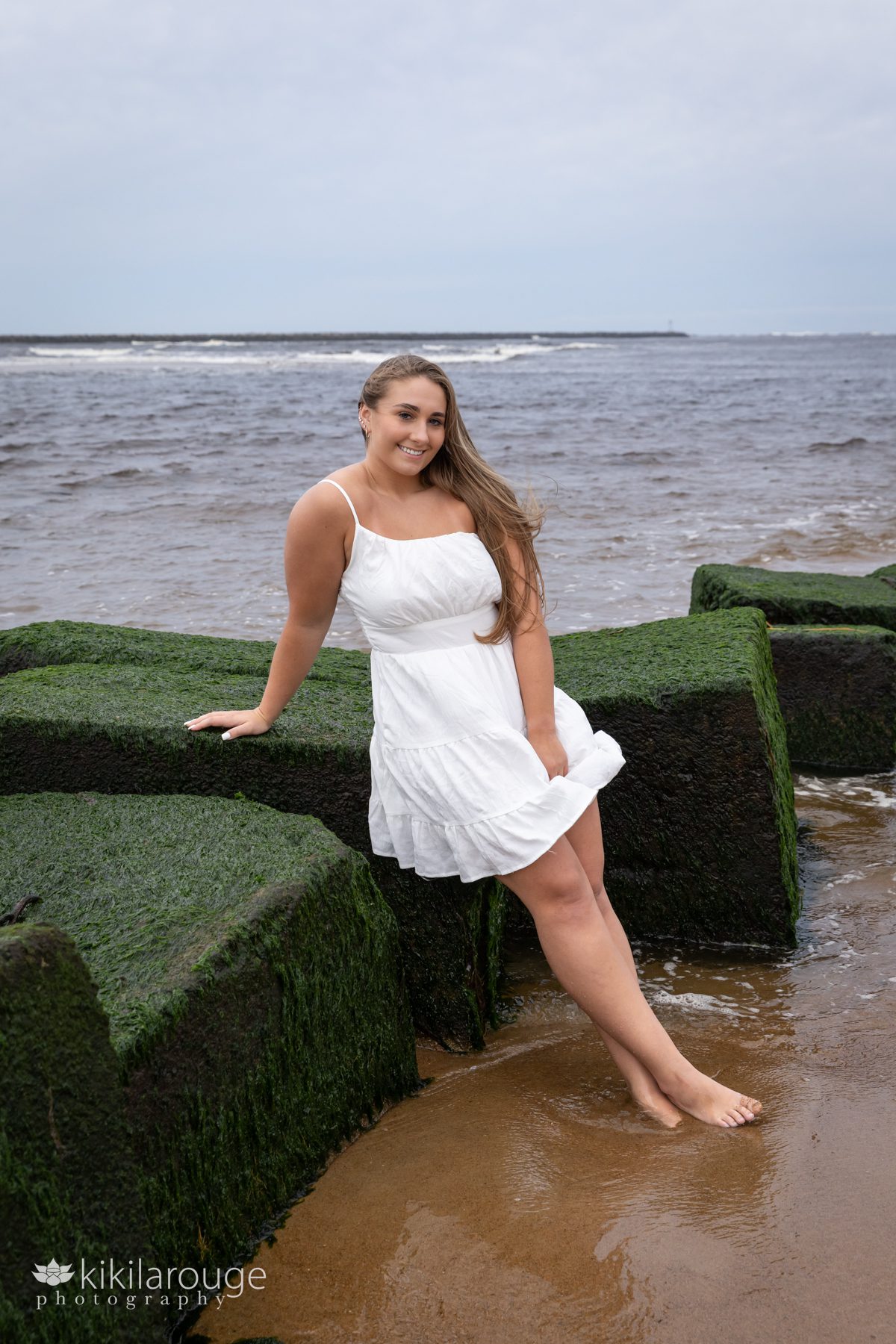 Triton Senior Portrait Girl in Jeans with white top tied in back at beach sitting on green seaweed covered blocks