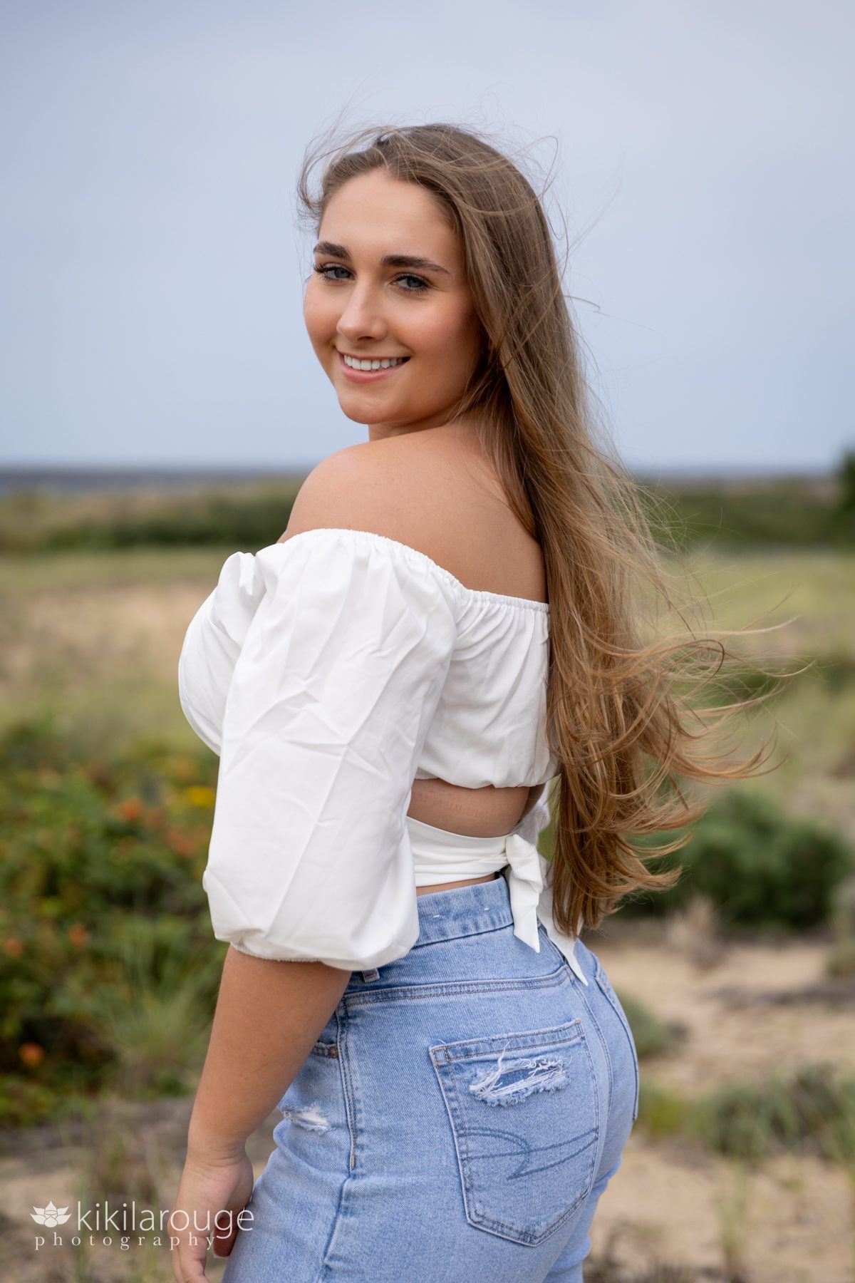 Triton Senior Portrait Girl in Jeans with white top tied in back at beach looking over shoulder