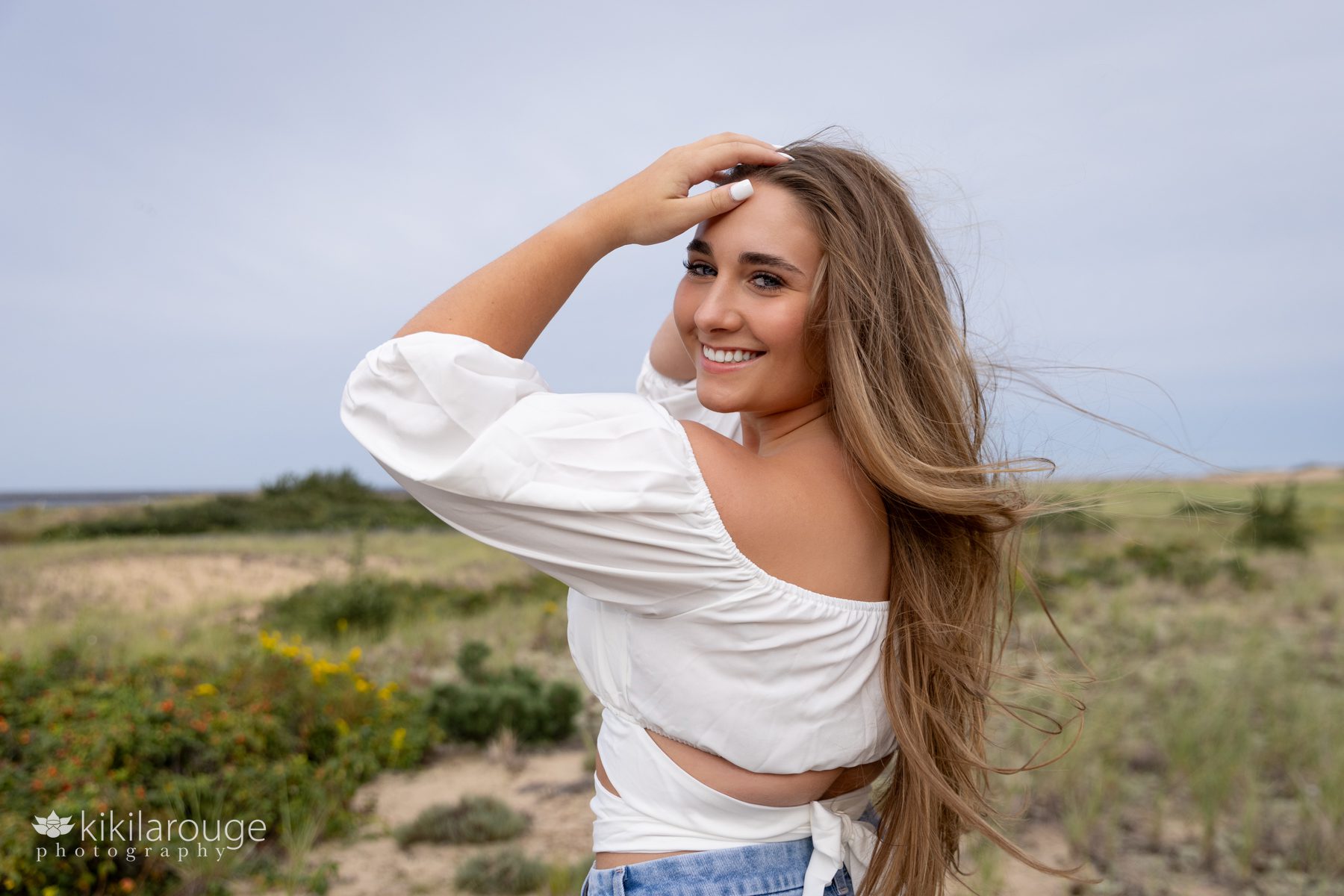 Triton Senior Portrait Girl in Jeans with white top tied in back at beach smiling with hands in air