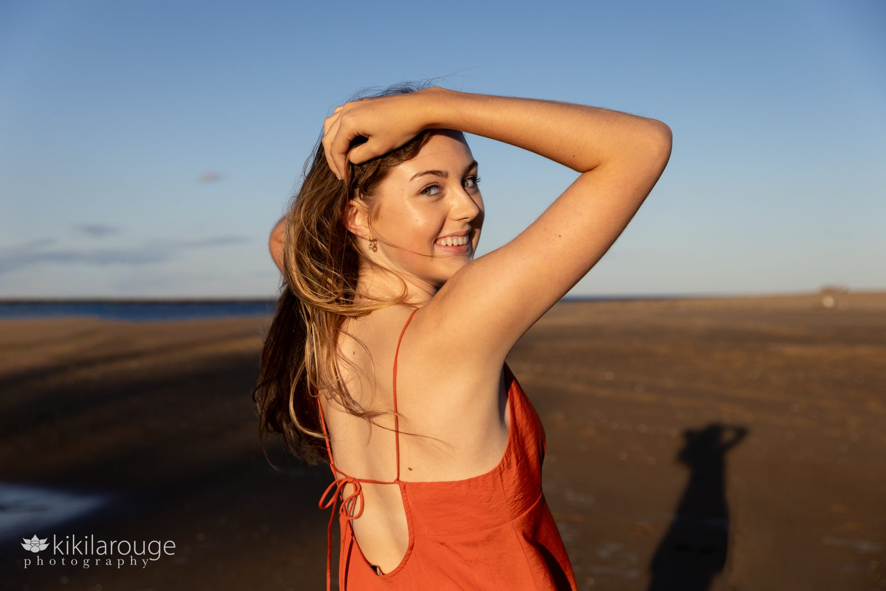 Teen girl in orange spaghetti strapped dress in bright sun at beach with hands in hair looking back through shoulder at camera