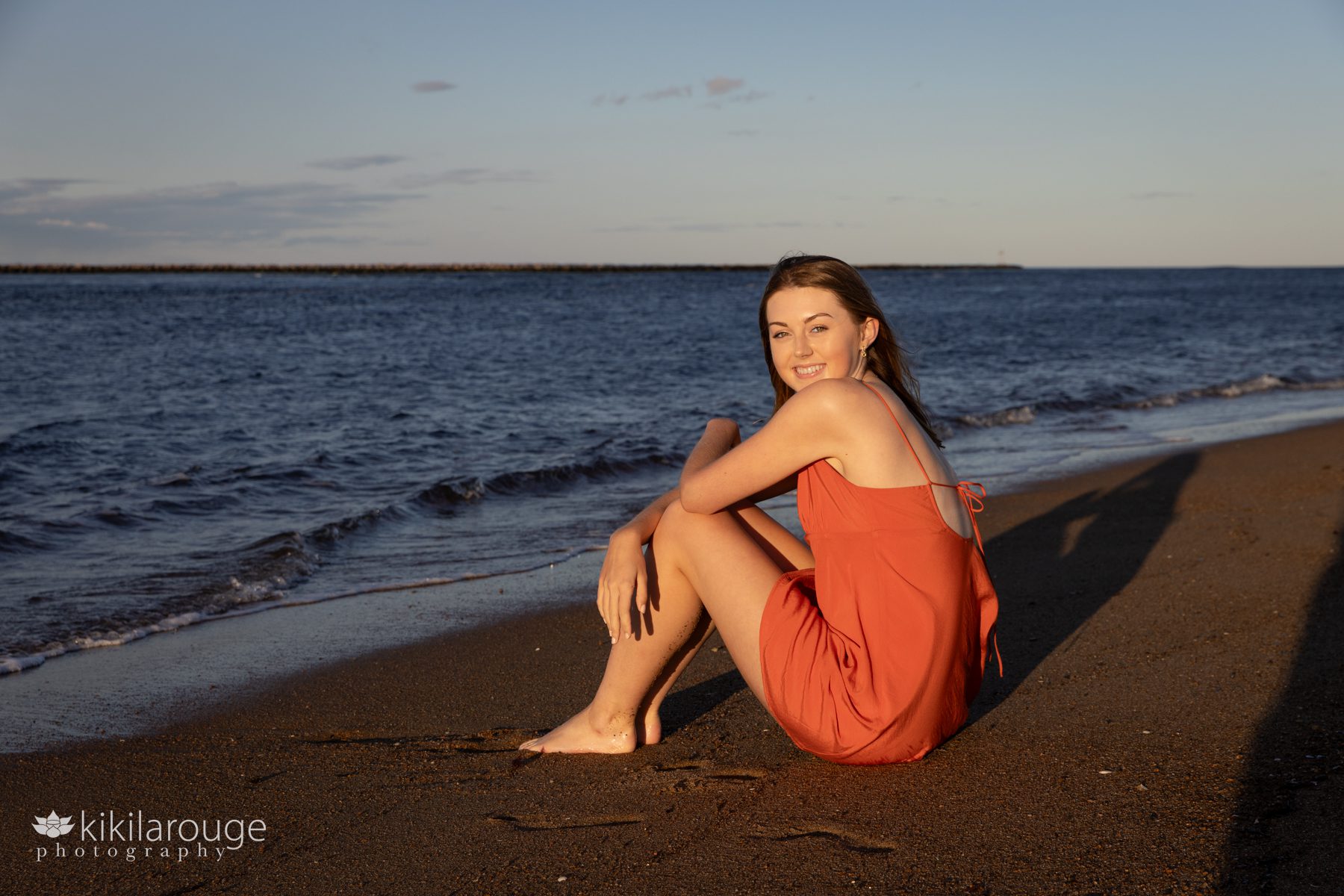 Teen girl in orange spaghetti strapped dress sitting at water's edge on Plum Island Beach at sunset