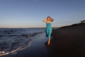 Teen girl in flowing blue dress walking along the water's edge at beach with hands in hair