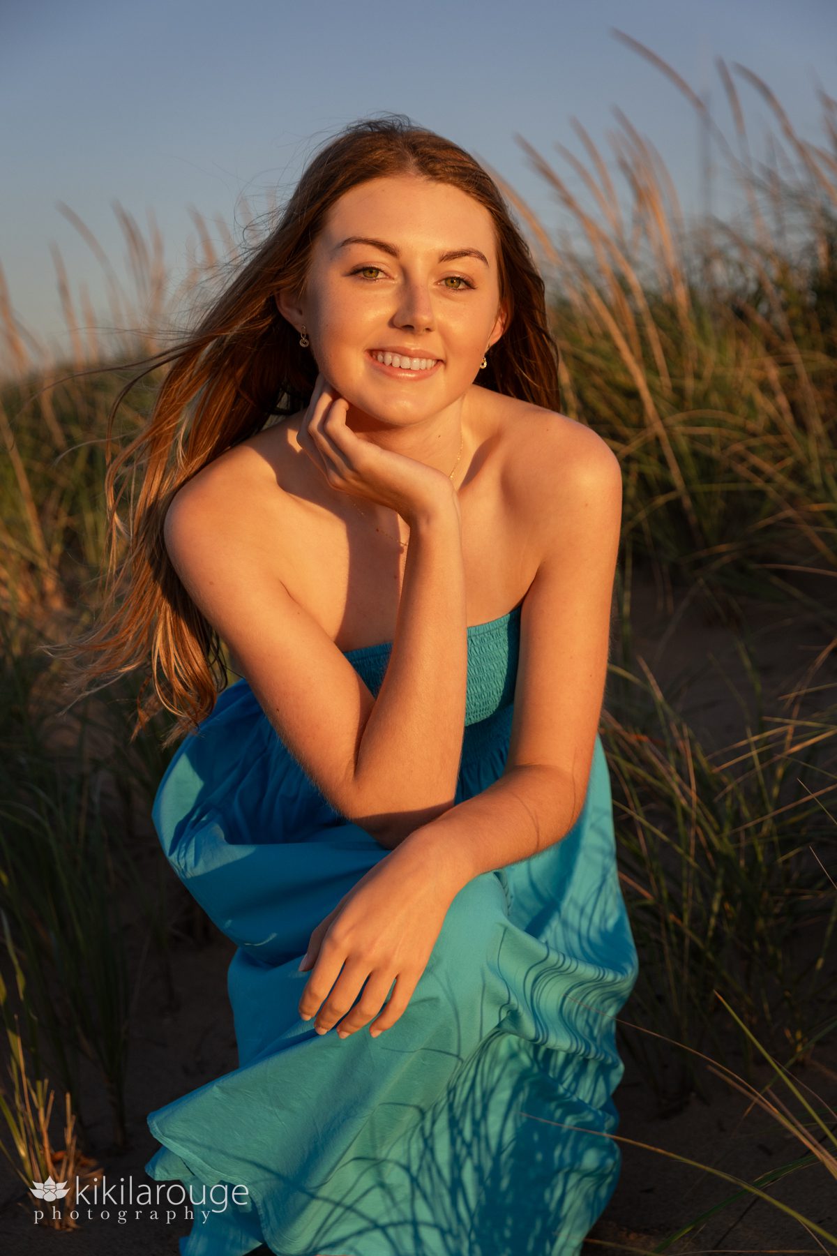 Teen girl from Masconomet Class of 2024 in blue dress sitting in beach dunes with hand on neck at Plum Island Beach Sunset