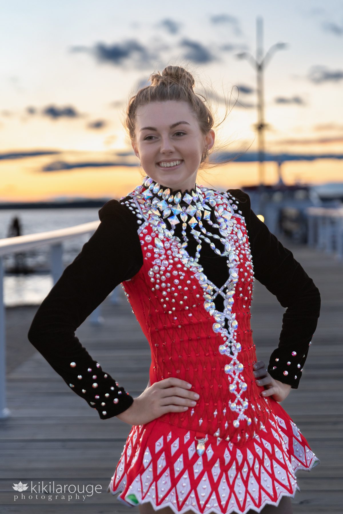 Girl smiling with dramatic sunset in backdrop with hands on hips in a sparkly red and white Irish Step Dancer outfit