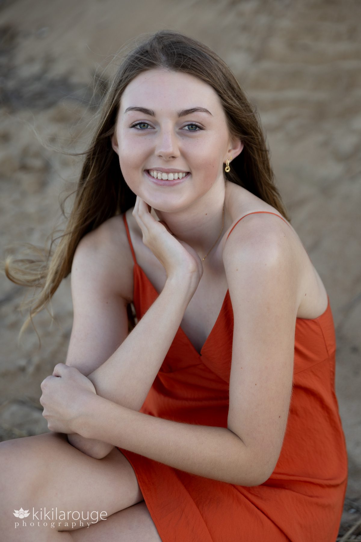 Teen girl in orange spaghetti strapped dress sitting in beach dune with hand under her neck