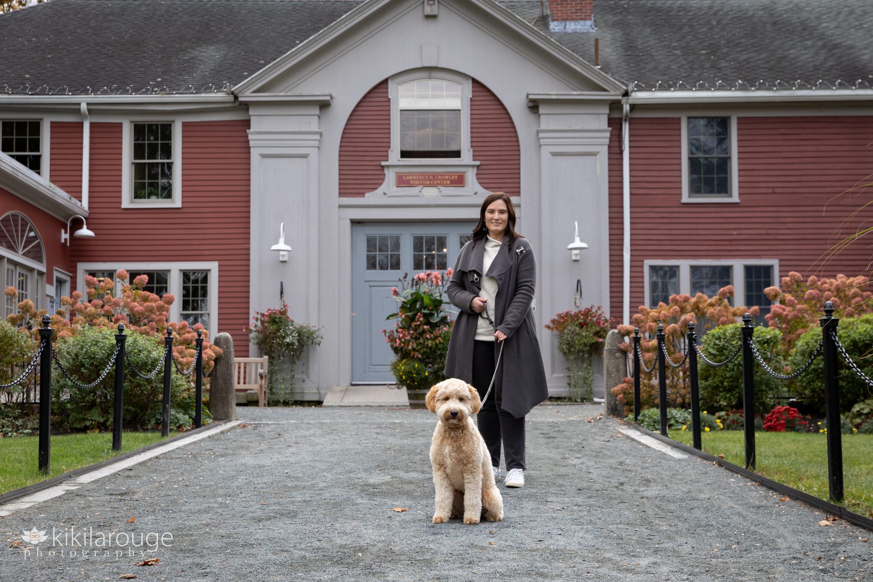 Woman in long gray coat in front of red building with labradoodle dog sitting in foreground fall imagery