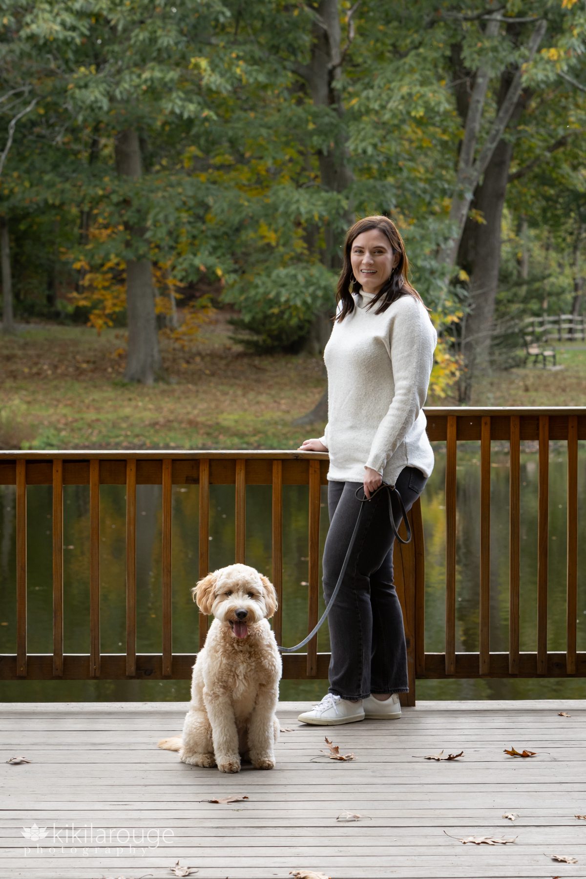 Woman in off white sweater and jeans on wood dock over looking pond with cute dog