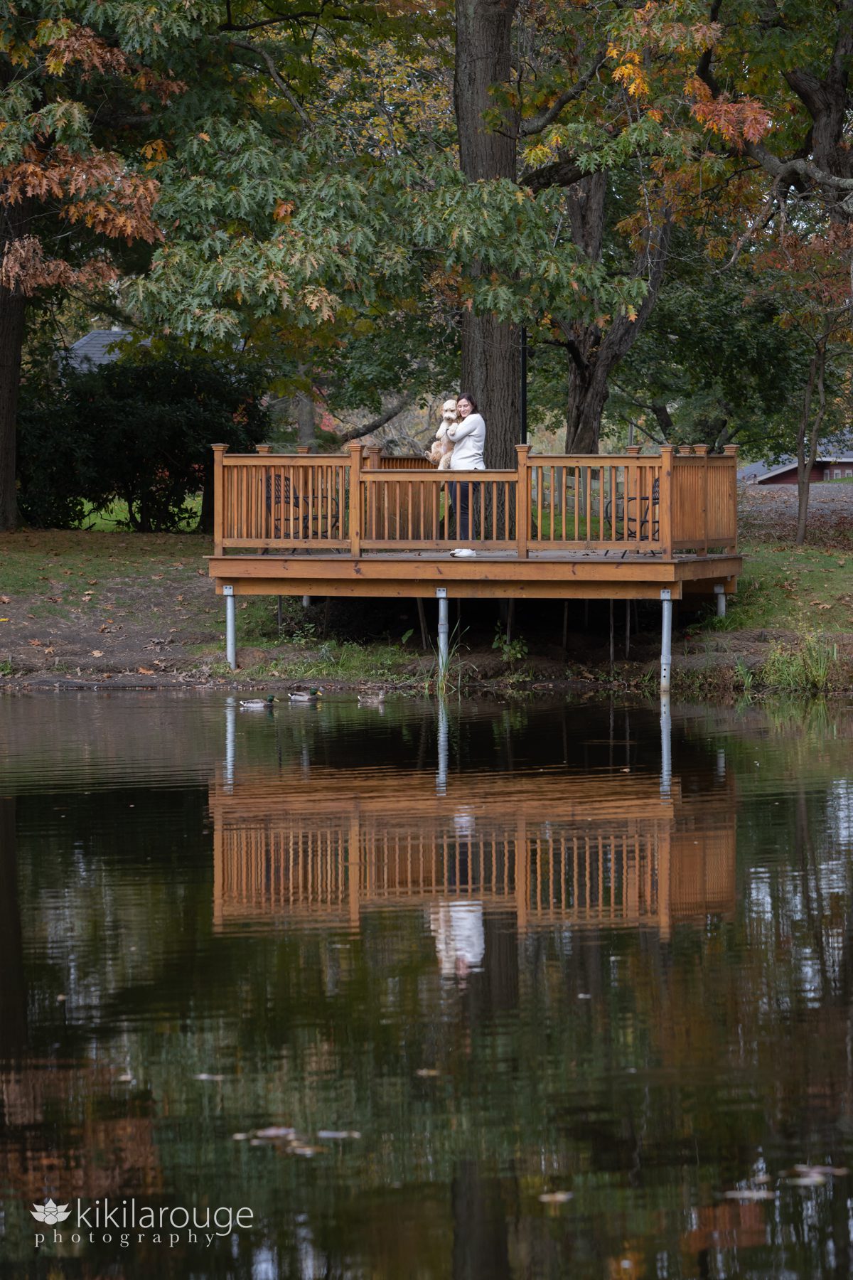 Woman holding her cute labradoodle pup on a wooden dock overlooking a pond with reflections of both