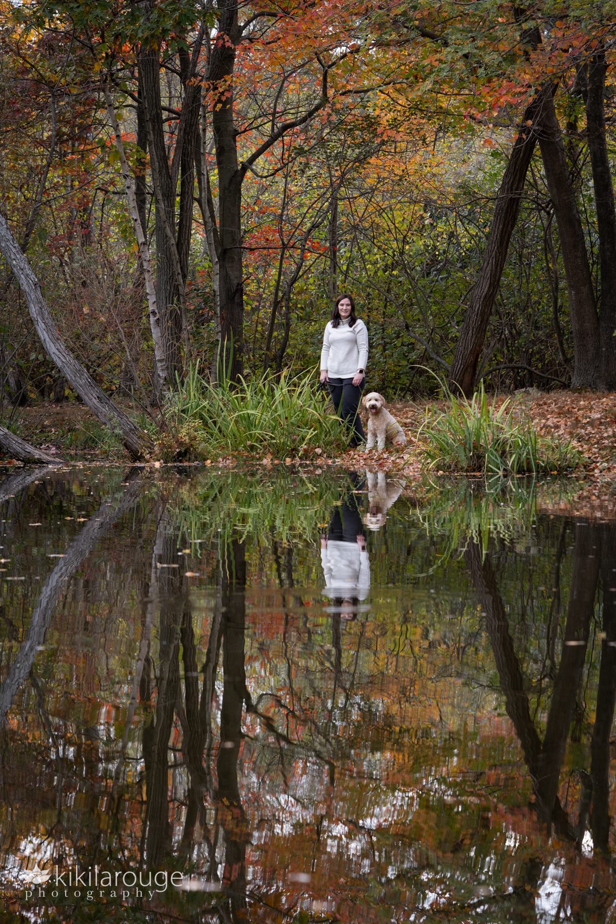Woman and dog at water's edge on a pond reflected with the foliage and trees surrounding them