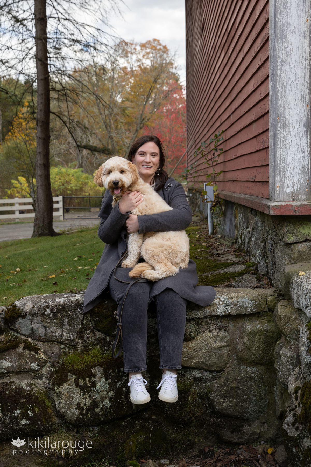 Woman in gray with long brunette hair sitting on stone wall hugging her cute labradoodle pup