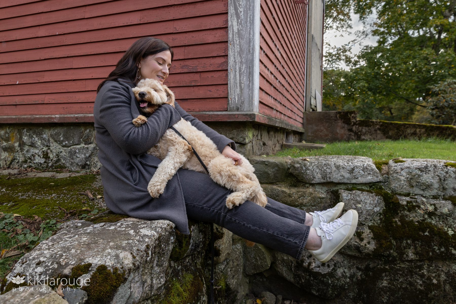 Woman in gray with long brunette hair sitting on stone wall hugging her cute labradoodle pup and snuggling