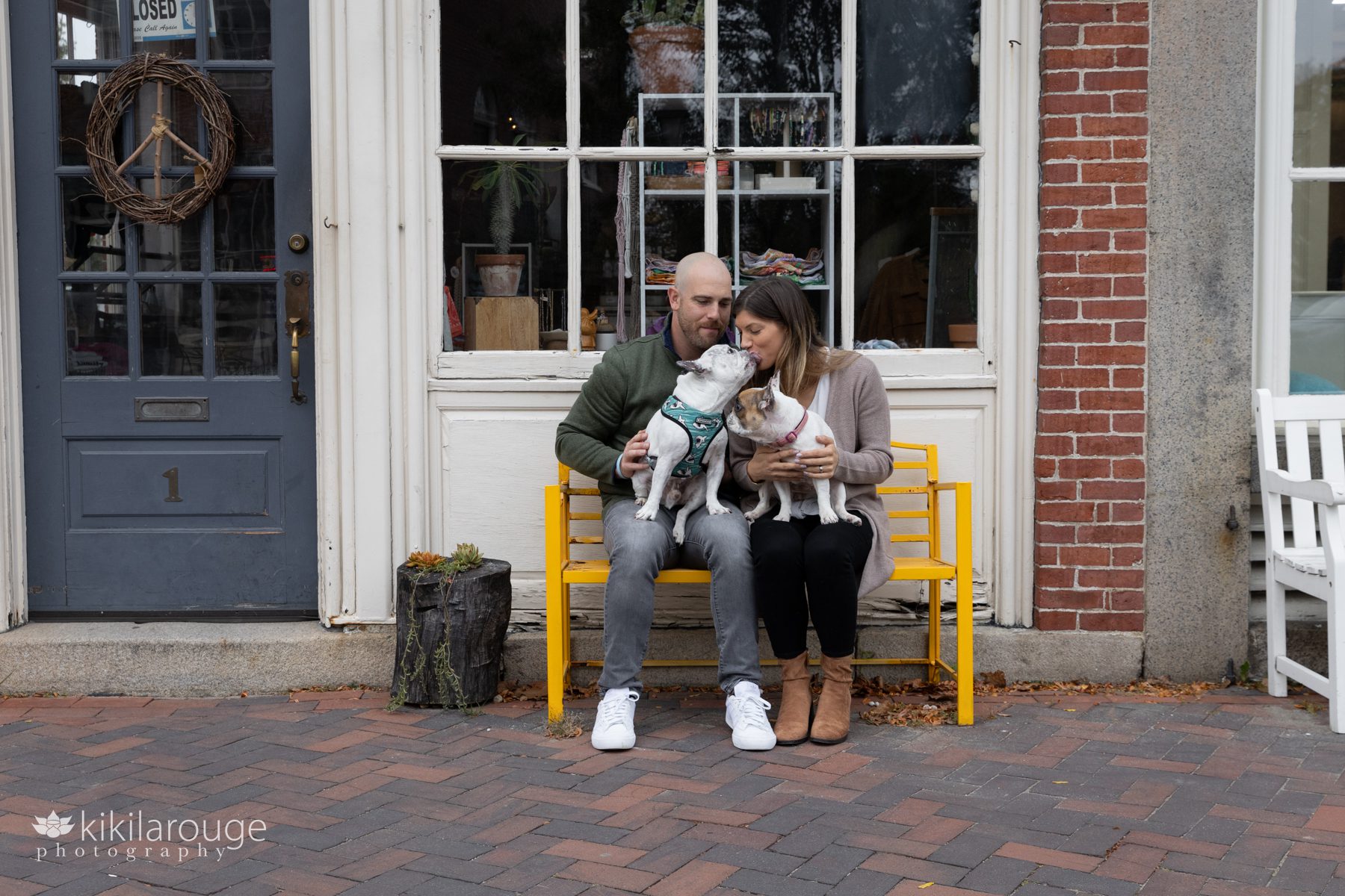 Couple sitting on yellow bench in front of story with their two white French bulldogs on their laps - one kissing the woman