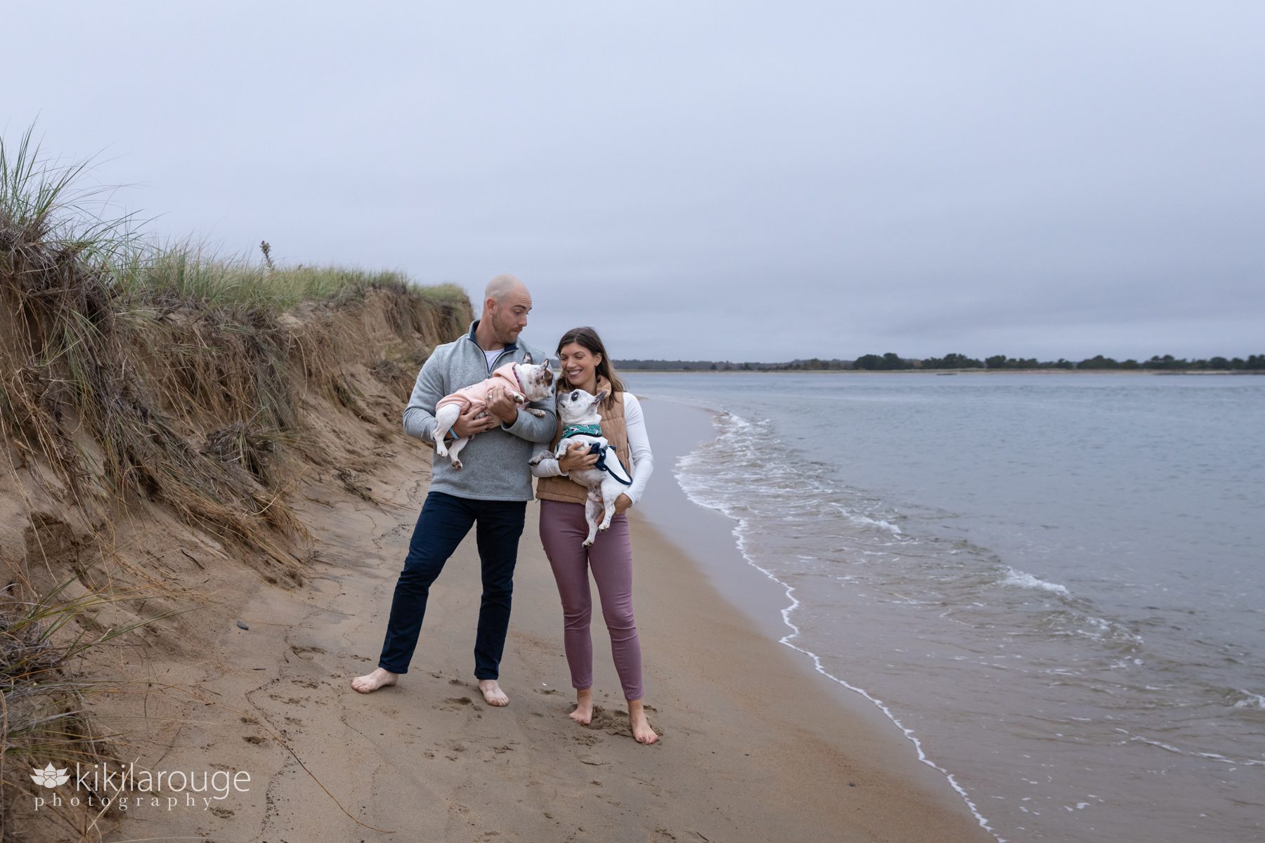 Cute outtake of couple smiling with their two French Bulldogs at the water's edge at Plum Island beach in Newburyport