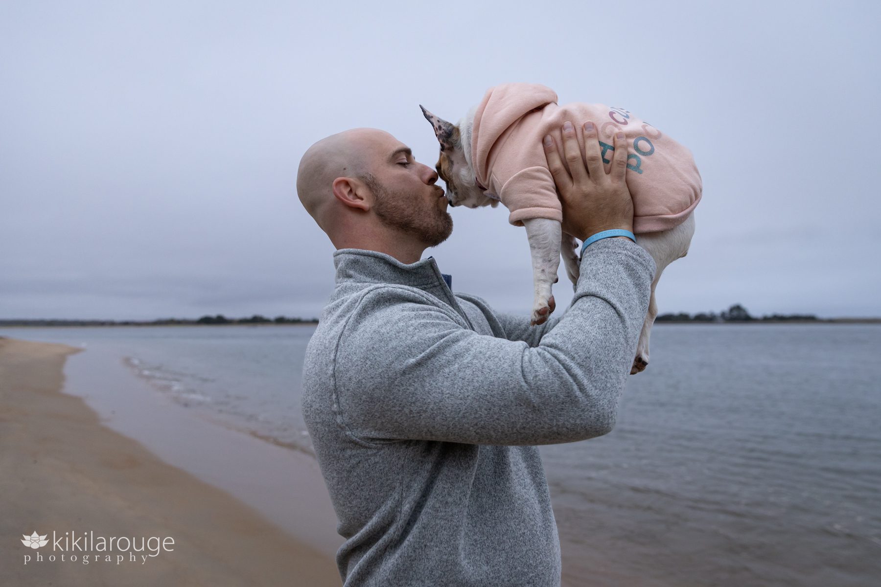 Bald man with red beard in gray sweater holding up tiny French bulldog in a Hocus Pocus sweatshirt at the beach Plum Island