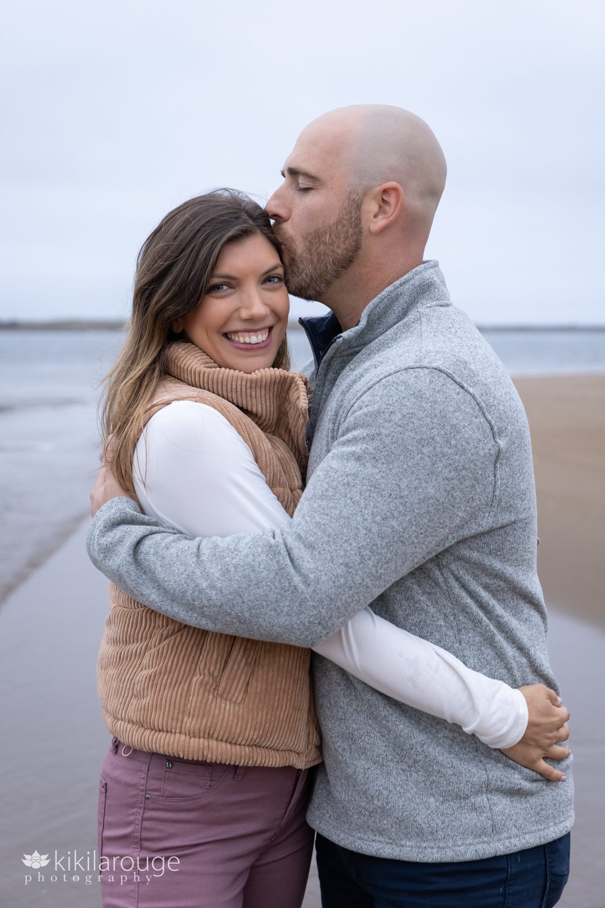 Man in gray sweater hugging and kissing wife on forehead at beach