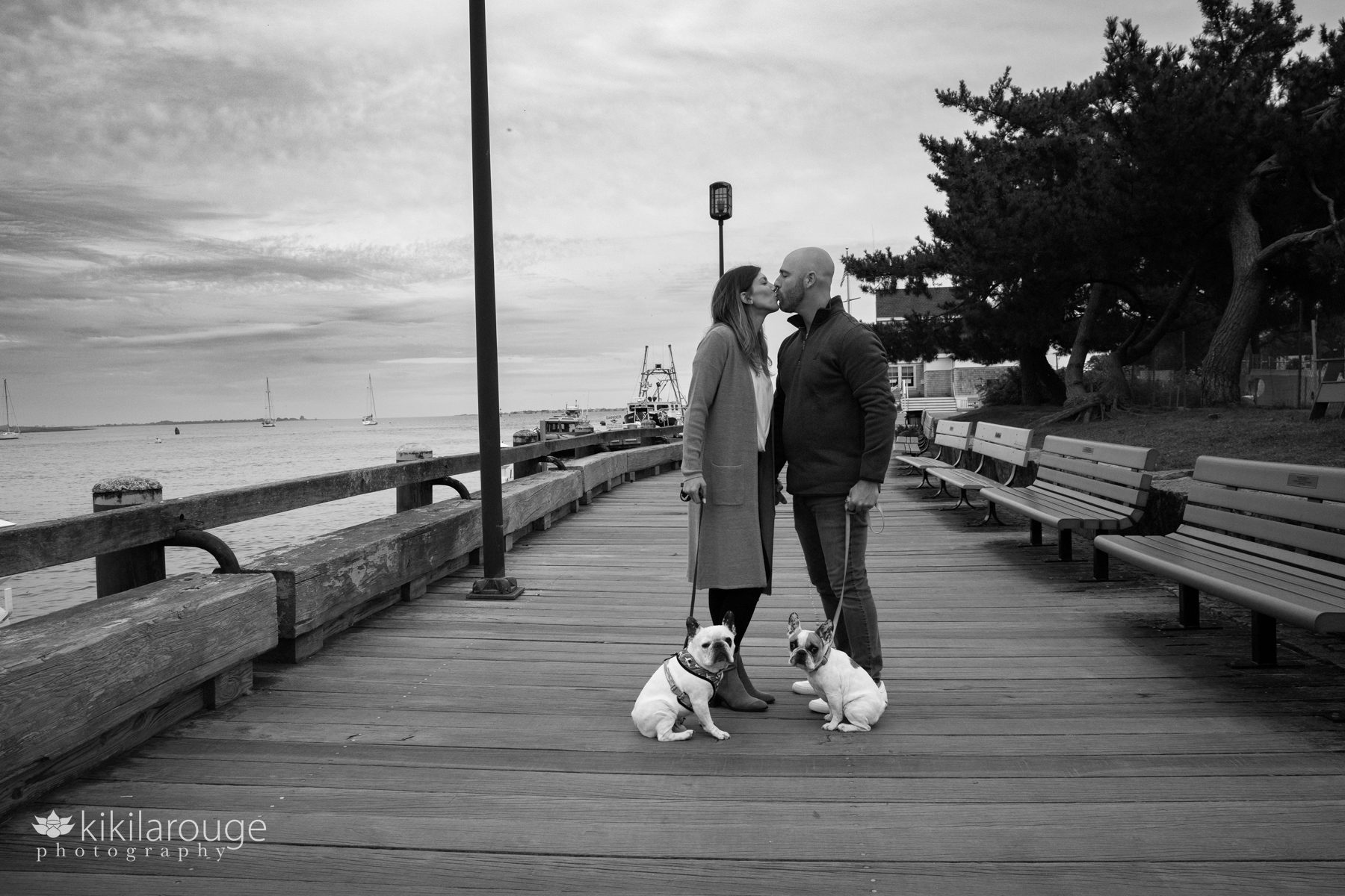 Couple kissing on Newburyport Waterfront Boardwalk with their two little French Bulldogs in tow