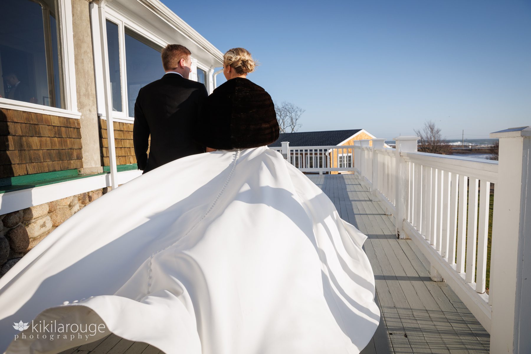 Wind blowing train of wedding dress up high as couple walks along a yacht club balcony