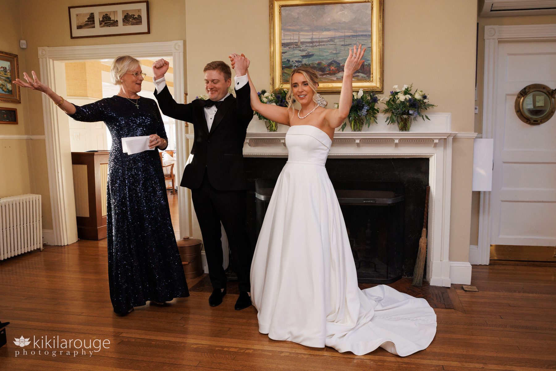 Couple and official with hands in air celebrating just getting married in yacht club in front of fireplace