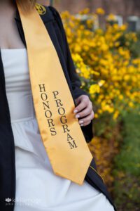 College graduate in cap and gown with yellow honors sash with yellow flowers in backdrop