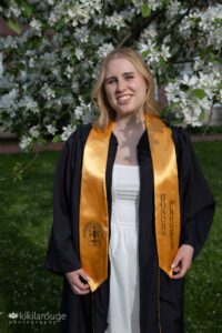 College graduate in cap and gown with yellow honors sash in sunlight by tree in bloom Framingham State